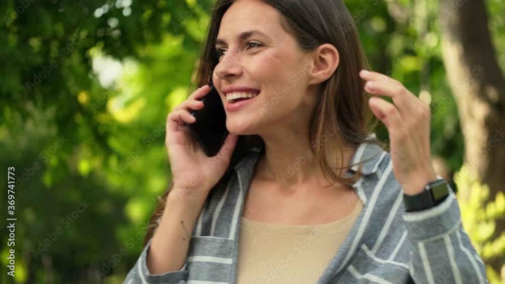 Smiling attractive brunette woman talking by smartphone and looking around in park Smiling attractive brunette woman talking by smartphone and looking around in park