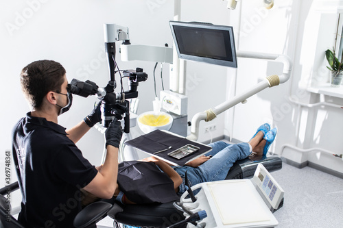 Portrait of young woman patient lying on dentist chair with open mouth. Young female dentist treating caries using microscope at the dentist office. Dentist wearing mask and gloves.
