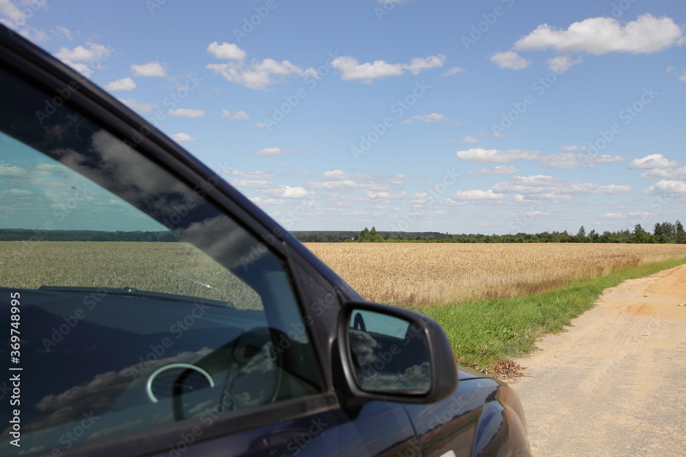 Car side door glass and mirror on Golden field, blue sky with white clouds and rural sandy road beautiful background, eco car tourism in Europe countryside 