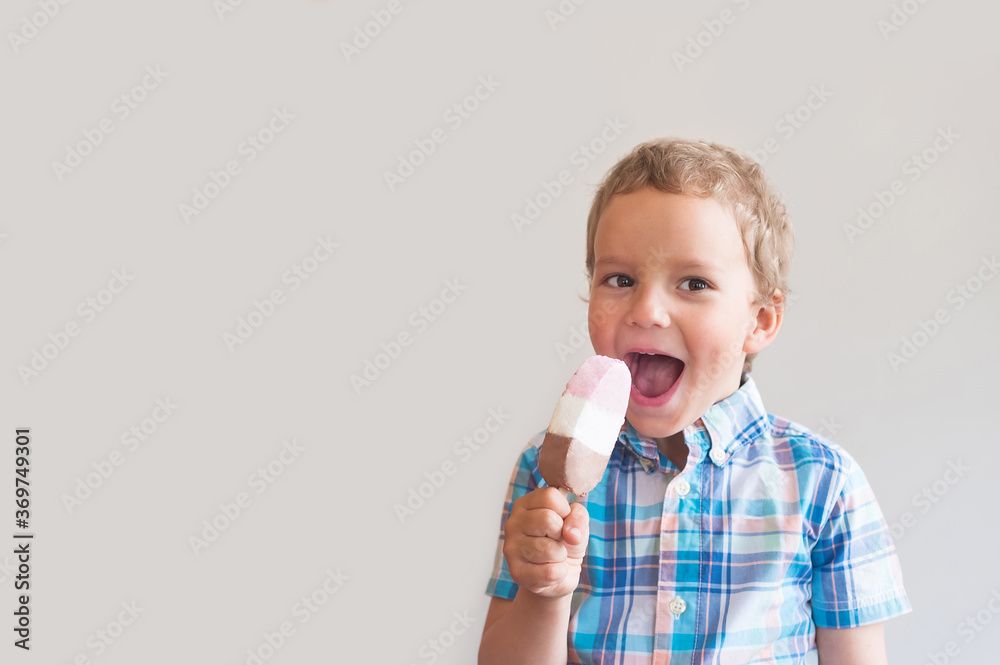 A beautiful fair-haired boy eats ice cream on a stick. Happy baby with ice cream against a gray background A beautiful fair-haired boy eats ice cream on a stick. Happy baby with ice cream against a gray background