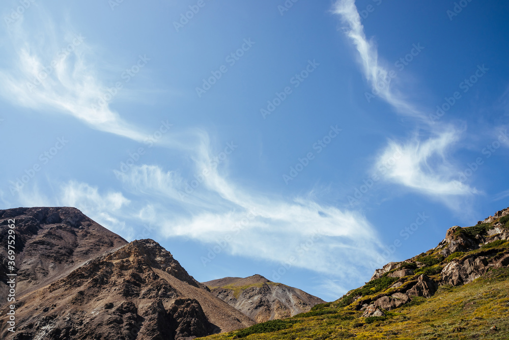 Scenic mountain landscape with beautiful cirrus clouds in clear blue sky over rocks in sunlight. Colorful highland scenery with spindrift clouds in blue clear sky above green brown rocky mountains.