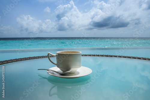 Coffee cup on a glass table with bright ocean and sky in Maldives.