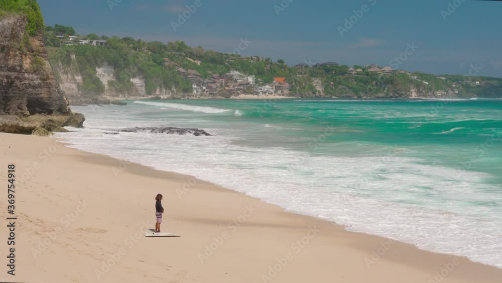 Surfer stand on Dreamland beach - popular surfing spot and looks out at the ocean with big waves and crystal clear sea water against the backdrop of picturesque nature island Bali, Indonesia. 4K Surfer stand on Dreamland beach - popular surfing spot and looks out at the ocean with big waves and crystal clear sea water against the backdrop of picturesque nature island Bali, Indonesia. 4K