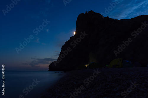 Tent on the beach at night. Full moon over the mountain. Outdoor recreation. Night seascape. Traveling with tents. Summer vacation on the shore of the night sea.