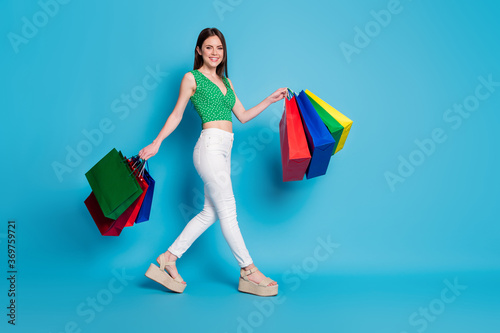 Full length profile side photo of cheerful girl hold bags go walk mall wear green crop tank-top dotted singlet isolated over blue color background