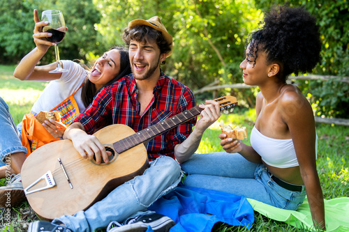 happy drunk multiracial friends laughing, singing and playing guitar drinking wine at a picnic in the countryside.