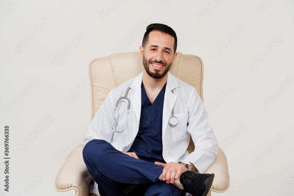 Arabian doctor surgeon in medical robe with phonendoscope seats and smile in armchair in studio on white blanked background. Confident arab on white background Arabian doctor surgeon in medical robe with phonendoscope seats and smile in armchair in studio on white blanked background. Confident arab on white background