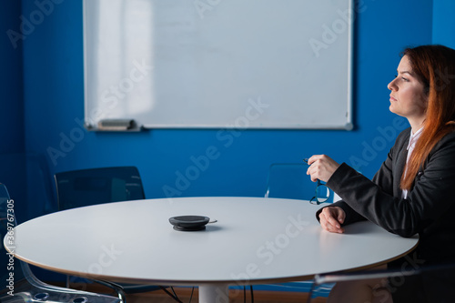 Pensive red-haired office manager in glasses and a suit in an empty conference room. Business woman waiting for important negotiations with partners and colleagues in the meeting room.