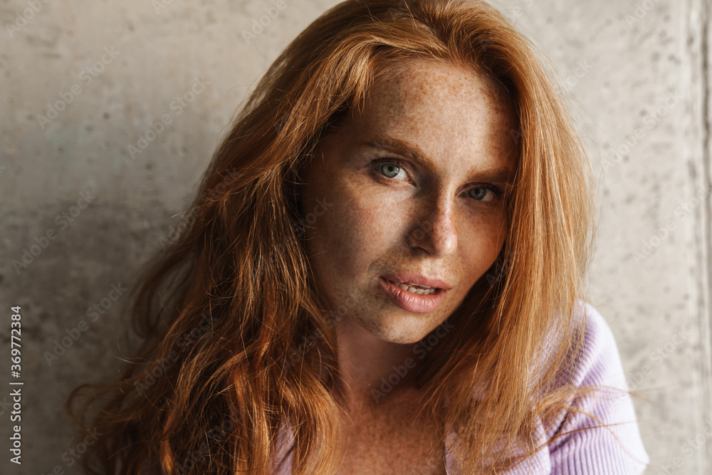 Image of redhead woman looking at camera while posing near concrete wall Image of redhead woman looking at camera while posing near concrete wall
