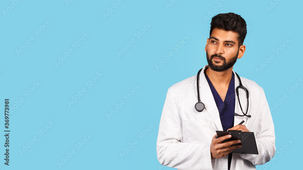 Handsome modern indian / asian doctor with stethoscope, in uniform writing prescription on tablet on blue background Handsome modern indian / asian doctor with stethoscope, in uniform writing prescription on tablet on blue background
