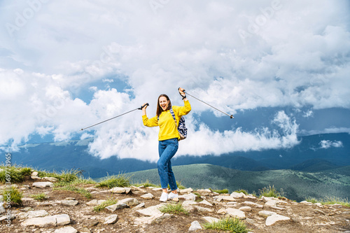Successful hiker woman standing with hands up on the mountain and holding sticks for nordic walk. A tourist girl achieved a top. Active lifestyle concept