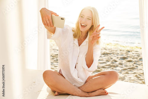 Image of woman gesturing peace sing while taking selfie on mobile phone