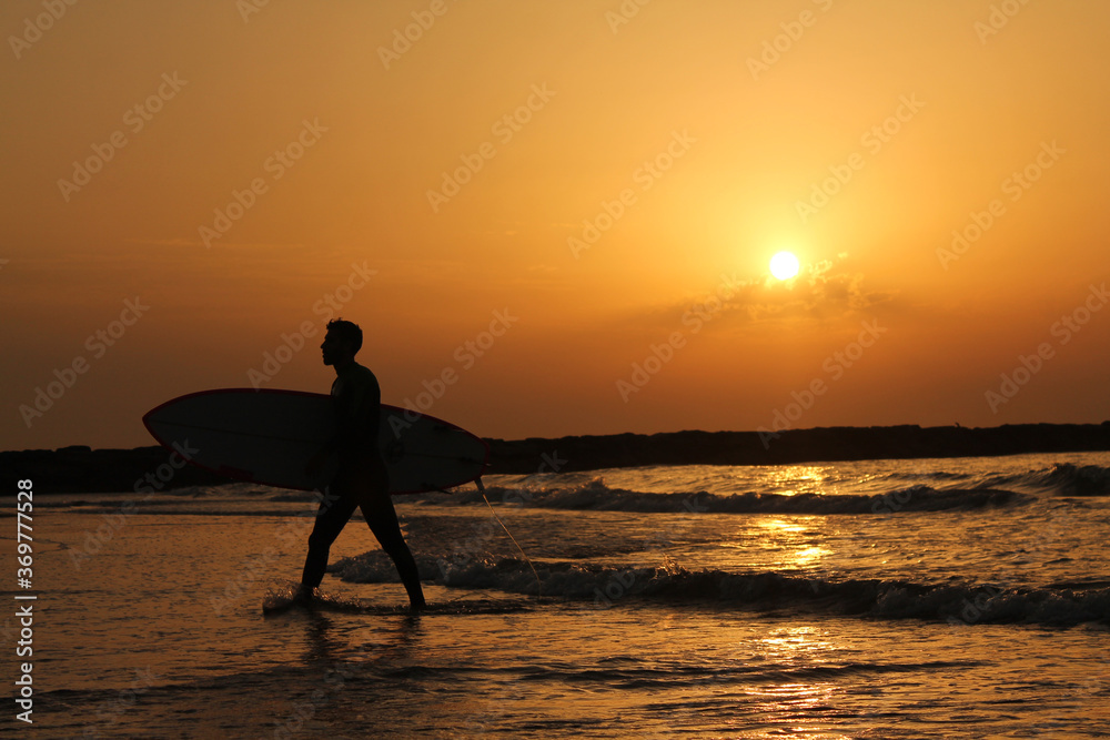 Surfer comes out of the water after playing sports. Unrecognizable man walking on the beach and enjoying of the beautiful sunrise.
