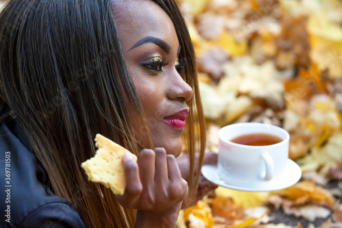 Beautiful young African girl eating cookies and drinking coffee from white Cup on autumn leaves background in Park