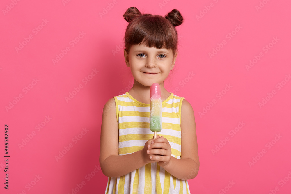 Little girl with big fruit ice cream standing against rosy wall and looking directly at camera, wearing summer outfit, having two knots, expressing positive emotions.