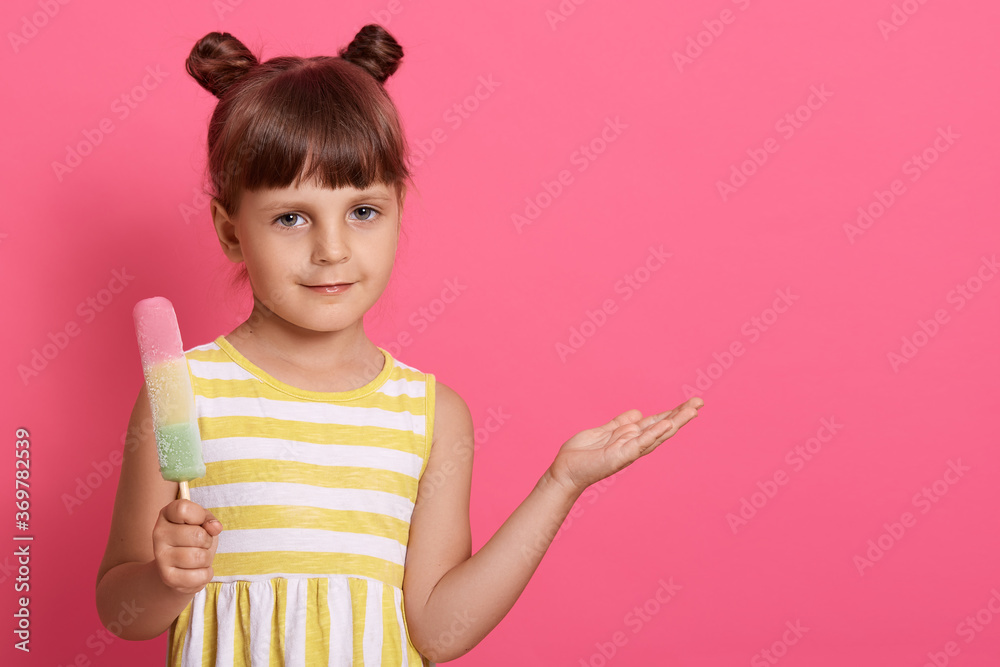 Cute female wearing summer white and yellow dress, having two knots, spreading palm aside and showing copy space, looks at camera, holding water ice. Cute female wearing summer white and yellow dress, having two knots, spreading palm aside and showing copy space, looks at camera, holding water ice.