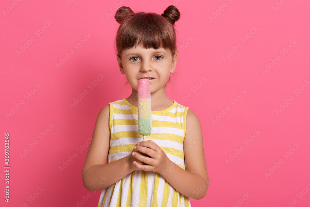 Cute child eats ice cream and expressing positive emotions, wearing white and yellow dress and having two knots, posing isolated over pink background. Cute child eats ice cream and expressing positive emotions, wearing white and yellow dress and having two knots, posing isolated over pink background.