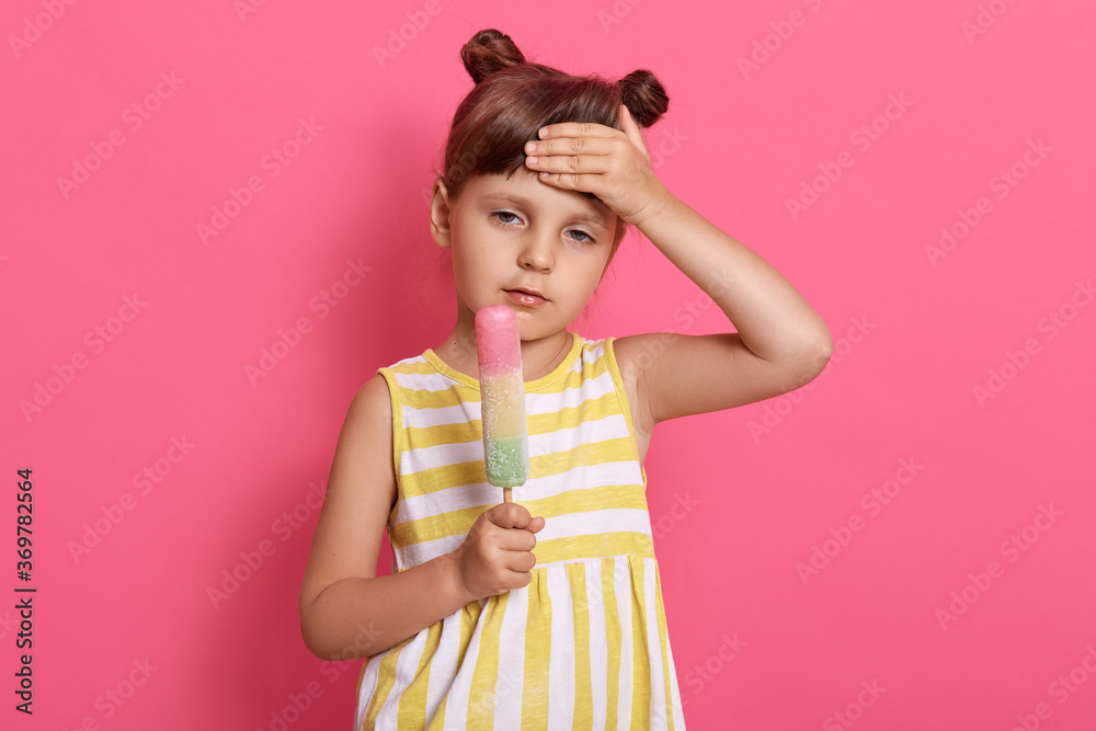 Charming girl holding ice cream and touching her forehead, has headache and high temperature, child wearing summer dress, posing isolated over pink background. Charming girl holding ice cream and touching her forehead, has headache and high temperature, child wearing summer dress, posing isolated over pink background.