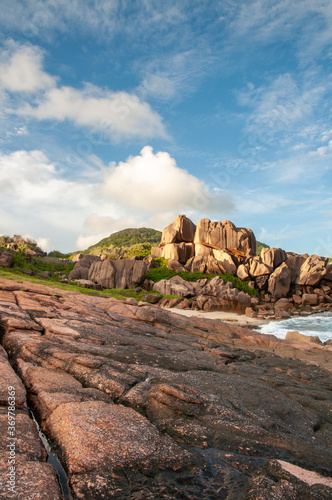Rough and wild rocky coastline at Anse Songe with lush green grass, tropical vegetation, red granite rocks on the waterfront of the Indian Ocean. La Digue island, Seychelles