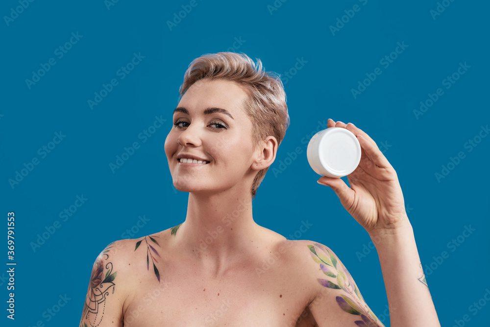 Portrait of beautiful half naked tattooed woman with short hair holding white plastic jar of cream or body lotion isolated over blue background Portrait of beautiful half naked tattooed woman with short hair holding white plastic jar of cream or body lotion isolated over blue background