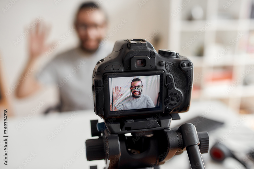 Young male blogger waving hand, saying hello, greeting subscribers while recording video blog or vlog at home studio. Focus on camera screen