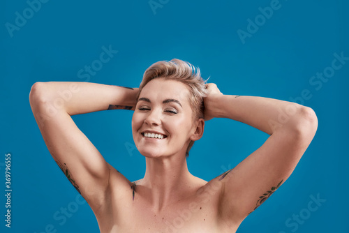 Beauty portrait of a young attractive half naked tattooed woman with perfect skin looking happy, arms folded behind her head isolated over blue background