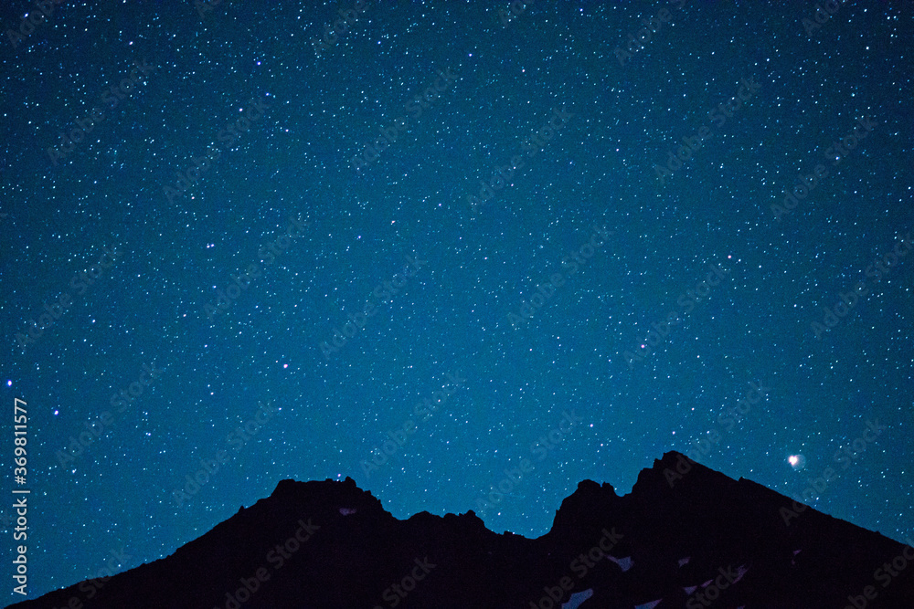 Stars dot the night sky above the silhouette of a broken volcanic mountain peak.