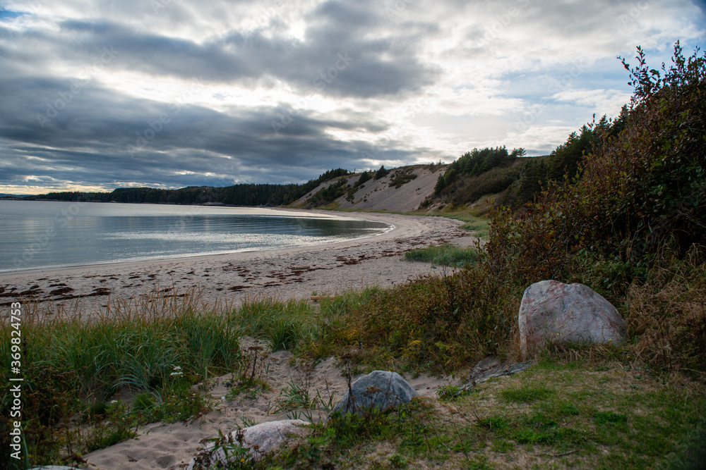 A sandy beach in a small cove with smooth ocean water. The sky is cloudy with the sun shining through. The foreground has large rocks, tall grass and shrubs. There are sandy mounds along the water. A sandy beach in a small cove with smooth ocean water. The sky is cloudy with the sun shining through. The foreground has large rocks, tall grass and shrubs. There are sandy mounds along the water.