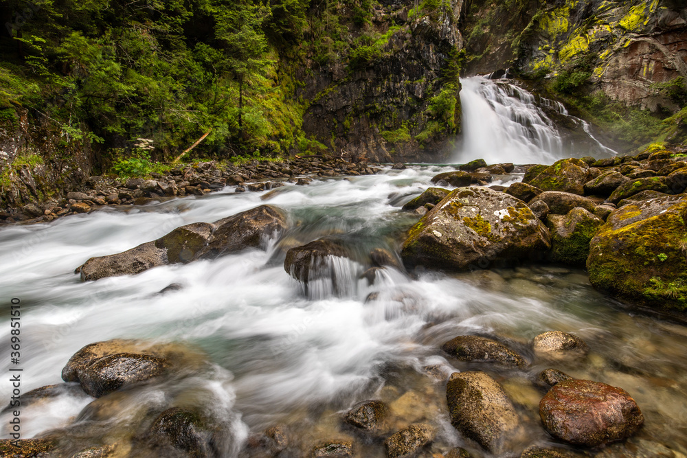 Fototapeta premium Wide angle view of an alpine waterfall flowing out of the rock and between boulders and pebbles, surrounded by luxuriant vegetation