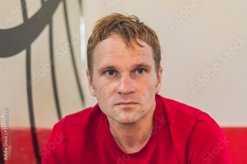 Serious sad handsome blond man in casual style red t-shirt sitting, waiting someone or changes, thinking. Skin without photoshop. Human relationship, difficulties during covid or life problems