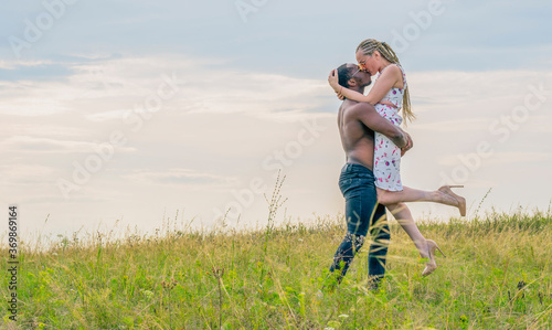 A dark-skinned young guy raised a white girl in his arms, and they froze in a gentle kiss against the backdrop of a green field and sunset
