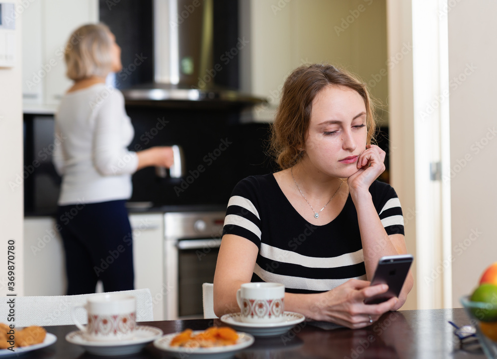 Pensive adult daughter using smartphone in kitchen interior while mature mother making tea