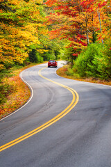A red car driving through winding road with beautiful autumn foliage trees in New England