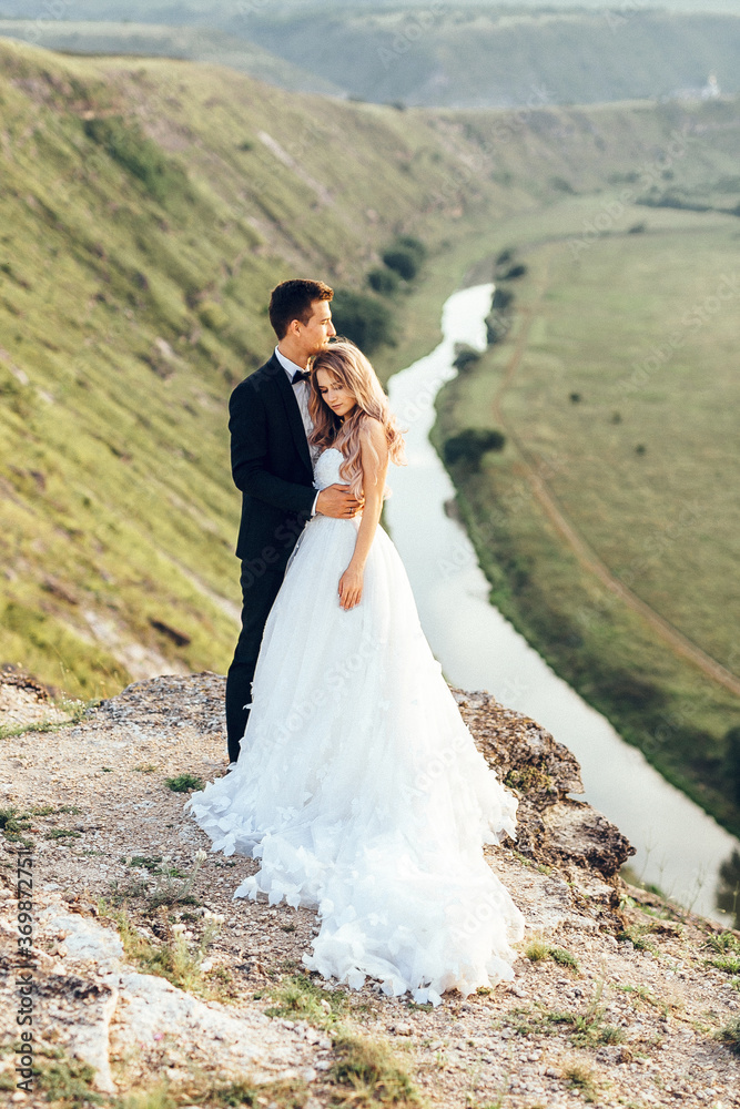 Full length body portrait of young bride and groom enjoying romantic moments outside at sunset in beautiful summer day. Wedding couple. Standing face to face with the green hills on background.