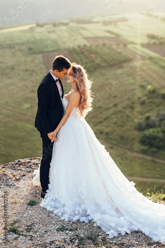 Full length body portrait of young bride and groom enjoying romantic moments outside at sunset in beautiful summer day. Wedding couple. Standing face to face with the green hills on background.