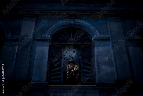 Halloween witch holding a skull standing in old damaged window with wall over cross  church  birds  dead tree and spooky cloudy sky  Halloween mystery concept