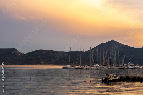 Fishing and sailing boats docked in Adamas Port on Milos Island, Greece during golden hour sunset, with calm flat sea and orange cloudy sky, mountains in the background. photo