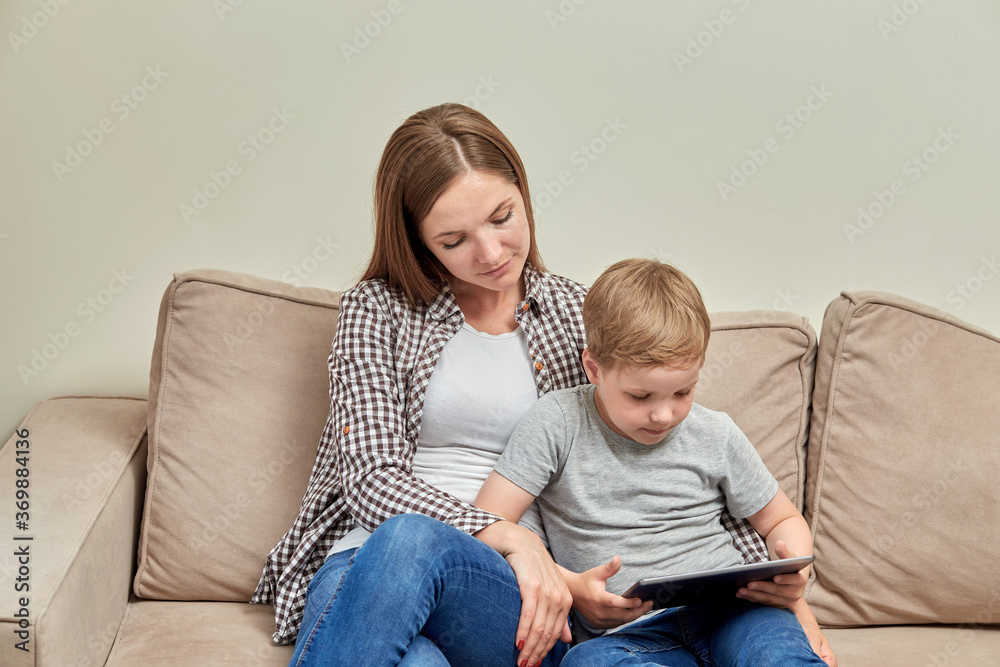 The son is playing in the tablet and his mother sits next to him and watches.