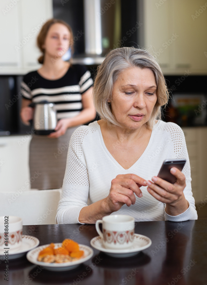 Positive senior woman using phone while young woman making tea