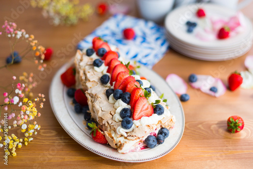 Homemade meringue roll with fresh berries - strawberries and blueberries - with porcelain tableware, napkins, flowers, berries and petals on wooden table