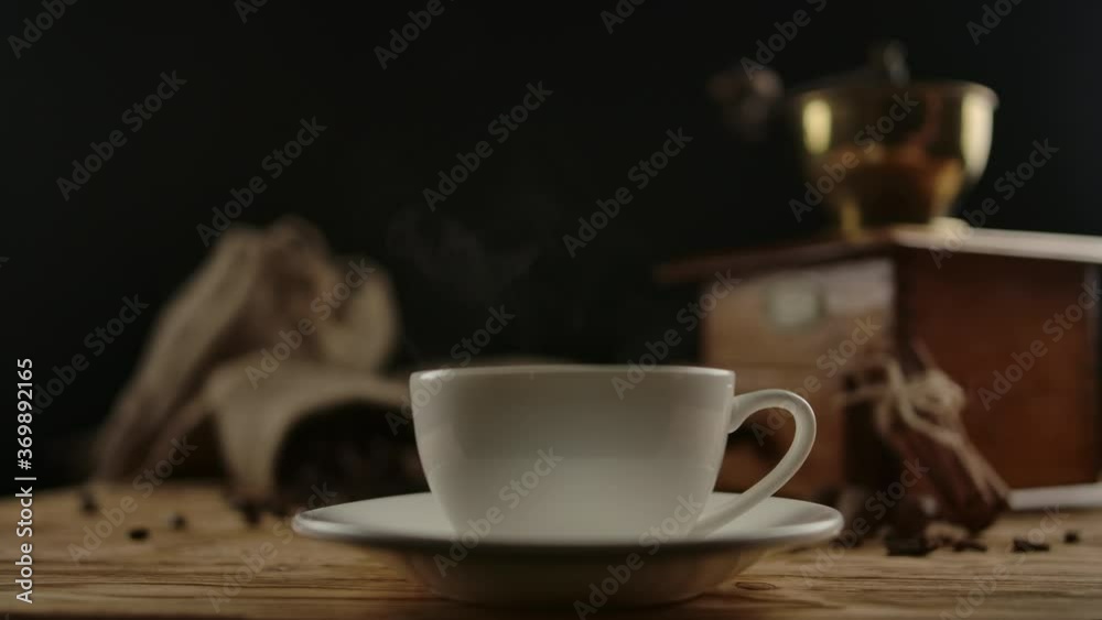 Serving coffee in elegant white porcelain cup by female hand. Traditional coffee break. Coffee beans, spices and manual grinder on background. Rotational shot.