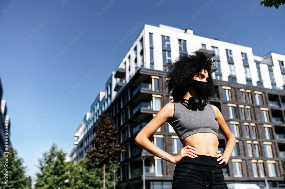 Sport in safety during quarantine. Close-up portrait of young beautiful multi-ethnic woman in sportswear with black medical mask on face in urban landscape. Woman looks away with hands on hips