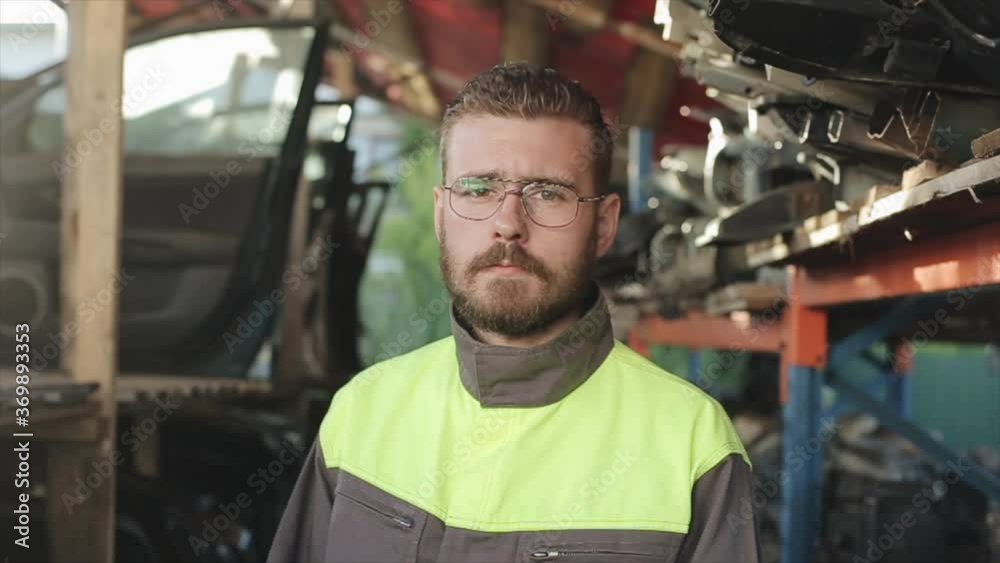 A young bearded auto mechanic wearing overalls stands in front of racks with spare parts. Close-up. Slow motion. The camera moves from left to right A young bearded auto mechanic wearing overalls stands in front of racks with spare parts. Close-up. Slow motion. The camera moves from left to right