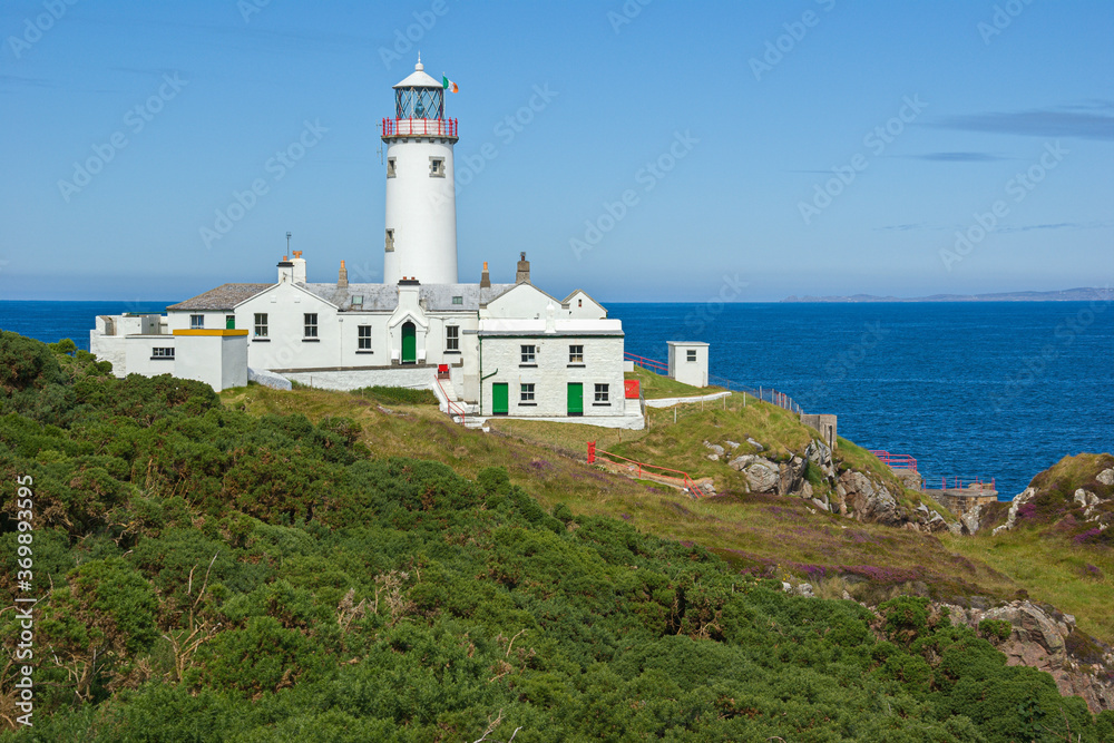 The white painted lighthouse at Fanad Head, Donegal, Ireland stands on a cliff top above the blue Atlantic Ocean, a safety beacon for shipping in the dangerous coastal waters around the rocky shores.