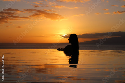  Silhouette of a girl standing in the pool, slightly turning her head. Against the background of the sunset and the ocean.