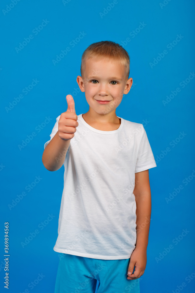 A young little Caucasian guy with blue eyes, dressed in a white t-shirt on a blue background, gives a thumbs up. A young little Caucasian guy with blue eyes, dressed in a white t-shirt on a blue background, gives a thumbs up.