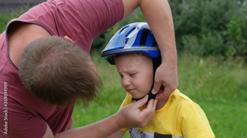 Father is putting safety helmet on his little son's head then teaching happy boy to ride bicycle