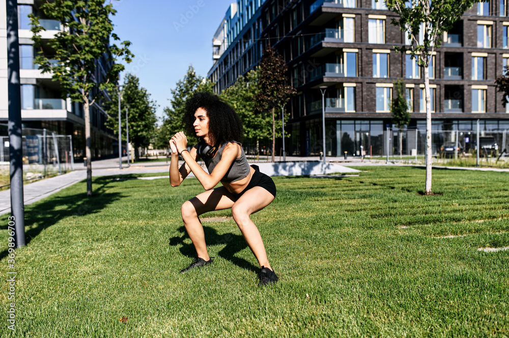 Fototapeta premium A young mixed-race woman doing fitness exercises outdoors. An African-American girl doing squats with an urban landscape on the background