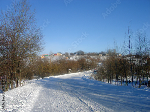 Sunny winter day. A winding road covered in snow goes up to the village. Trees with no leaves stand along the road