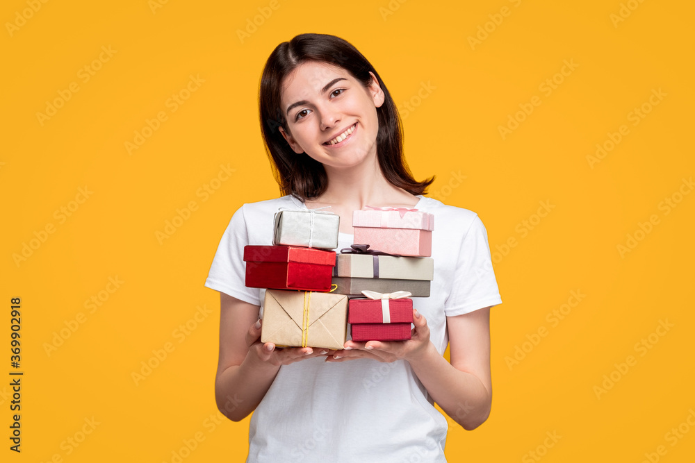 Holiday shopping. Discount sale. Cheerful female assistant in white t-shirt holding gift boxes pile isolated on orange background. Anniversary present. Happy birthday greeting. Holiday shopping. Discount sale. Cheerful female assistant in white t-shirt holding gift boxes pile isolated on orange background. Anniversary present. Happy birthday greeting.